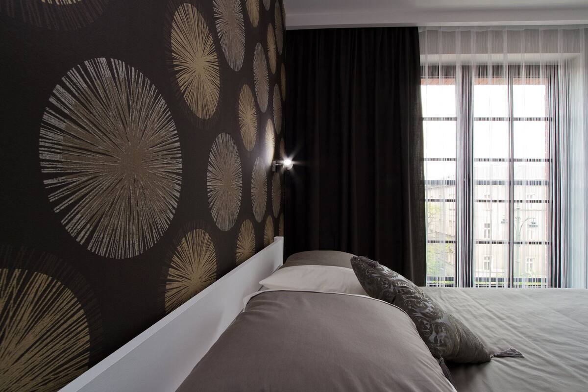 Ein modernes Hotelzimmer mit einem großen Bett, dunkel gemustertem Wandpapier und Blick auf die Stadt durch transparente Vorhänge.