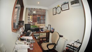 Un petit bureau avec un bureau, un ordinateur et une chaise. On y trouve des papiers, un téléphone et une fenêtre donnant sur des immeubles.