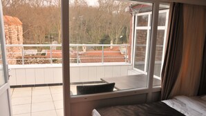 Une chambre avec un lit, une fenêtre donnant sur un immeuble et des arbres, et un balcon avec une balustrade.