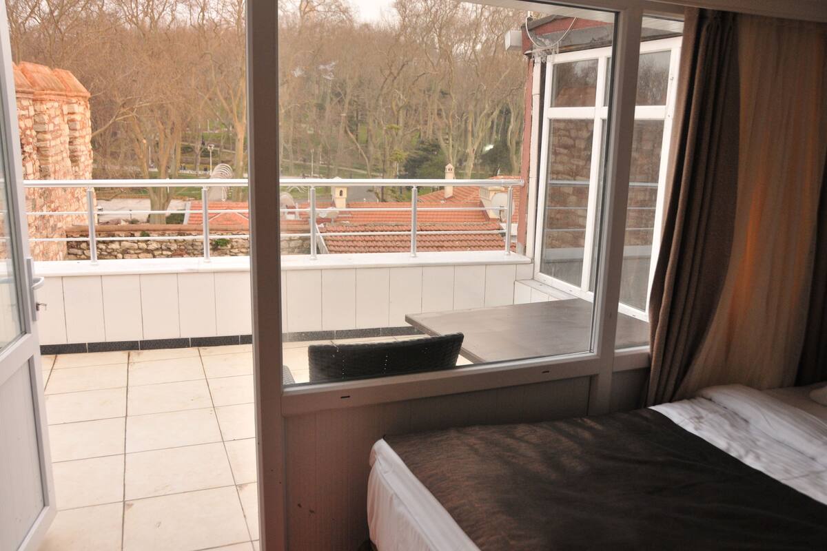 A room with a bed, a window with a view of a building and trees, and a balcony with a railing.