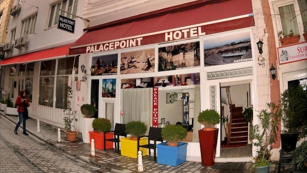 Un hôtel avec un auvent rouge, de nombreuses fenêtres et des places assises en plein air agrémentées de plantes en pot.