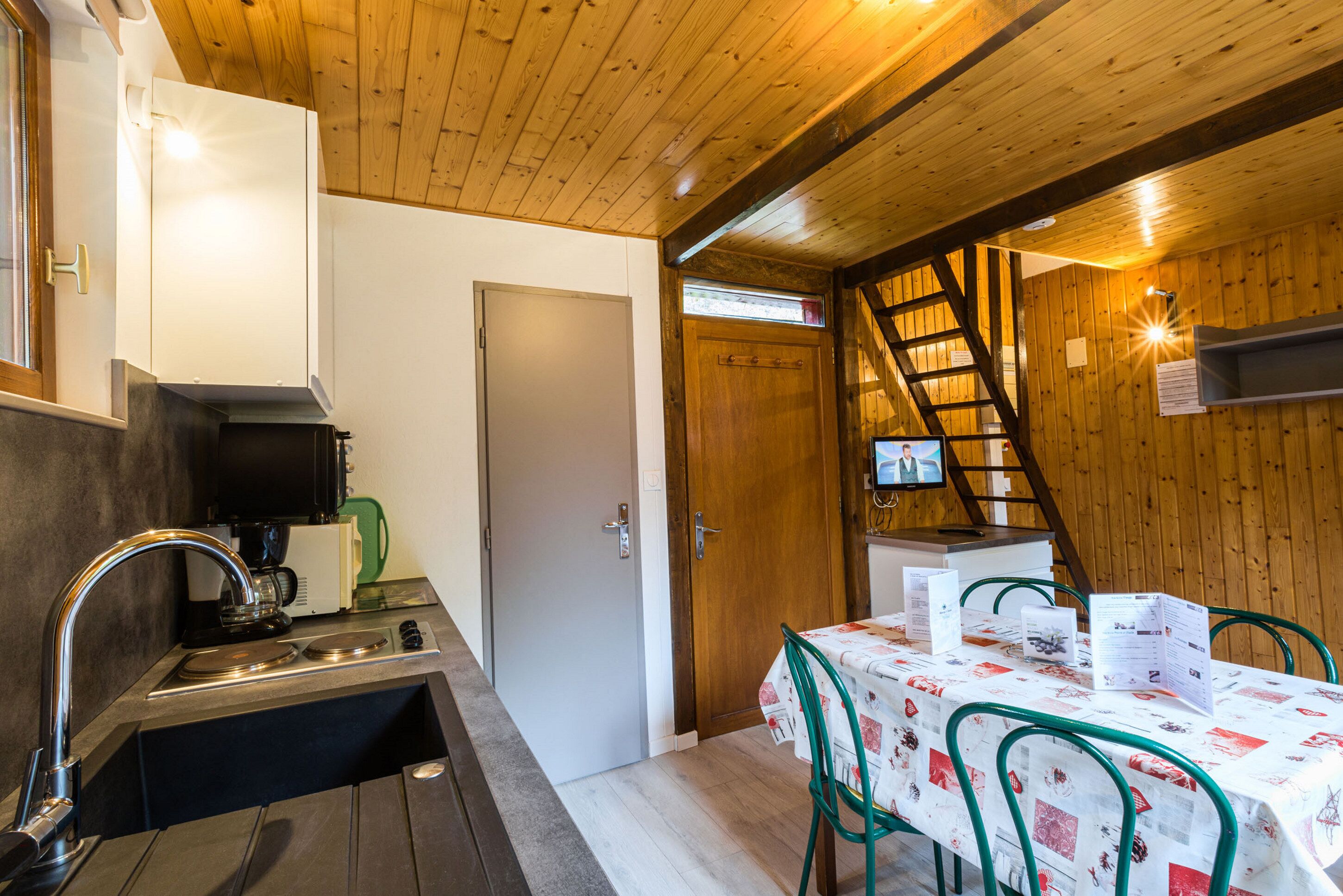 Una cocina compacta con lavabo, cocina y una cafetera pequeña. Una puerta de madera da a una escalera. Hay una mesa con sillas y un televisor montado en la pared.
