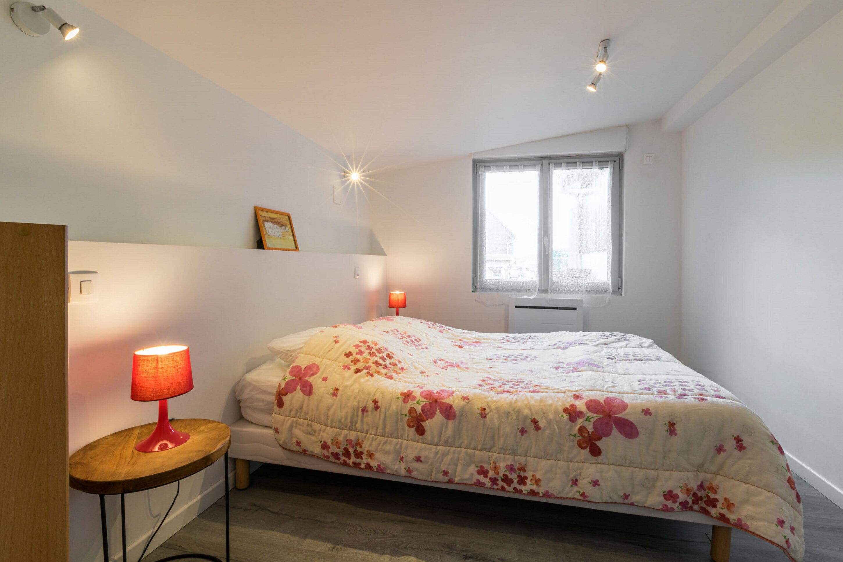 A bedroom with a bed, a nightstand, a red lamp, a window, and a ceiling light.