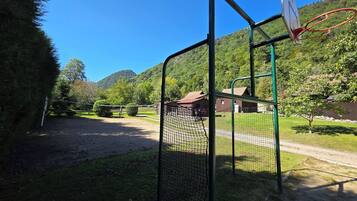 En basketballbane med net, omgivet af et hegn, med et hus og træer i baggrunden.