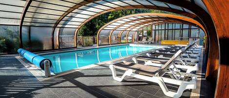 A modern indoor pool with a curved roof, lounge chairs, and a clear view of the surrounding greenery.