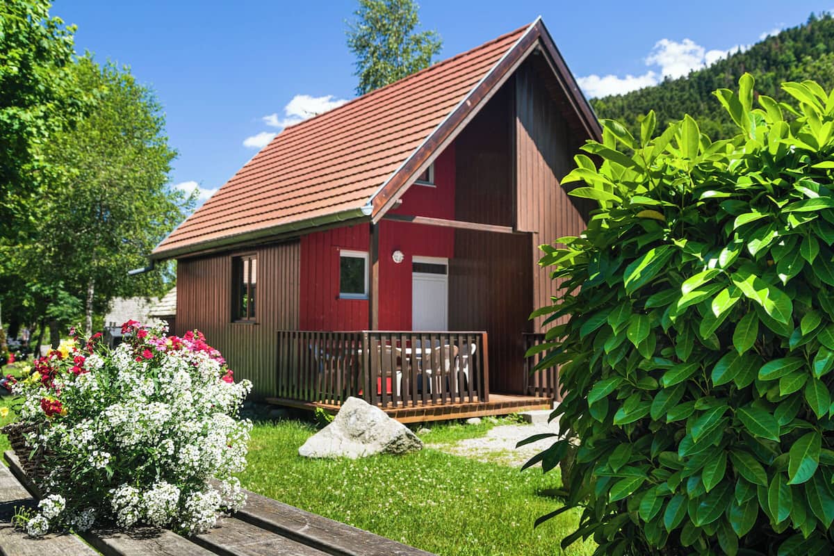 Una cabaña roja de madera con techo inclinado, un porche y un pequeño jardín con flores y una roca.