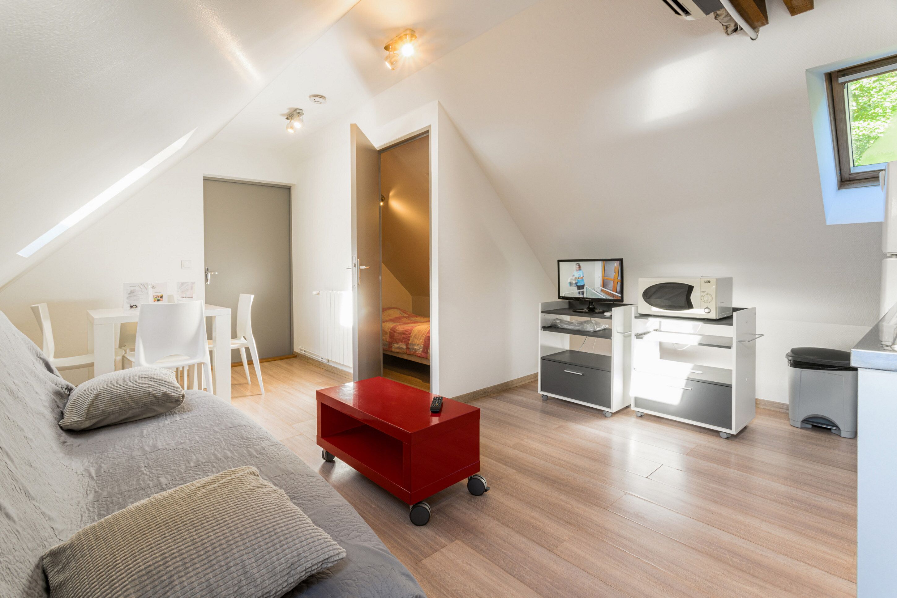 Une pièce moderne et bien éclairée, avec un lit en mezzanine, un bureau avec un ordinateur, une petite kitchenette et un chariot rouge.