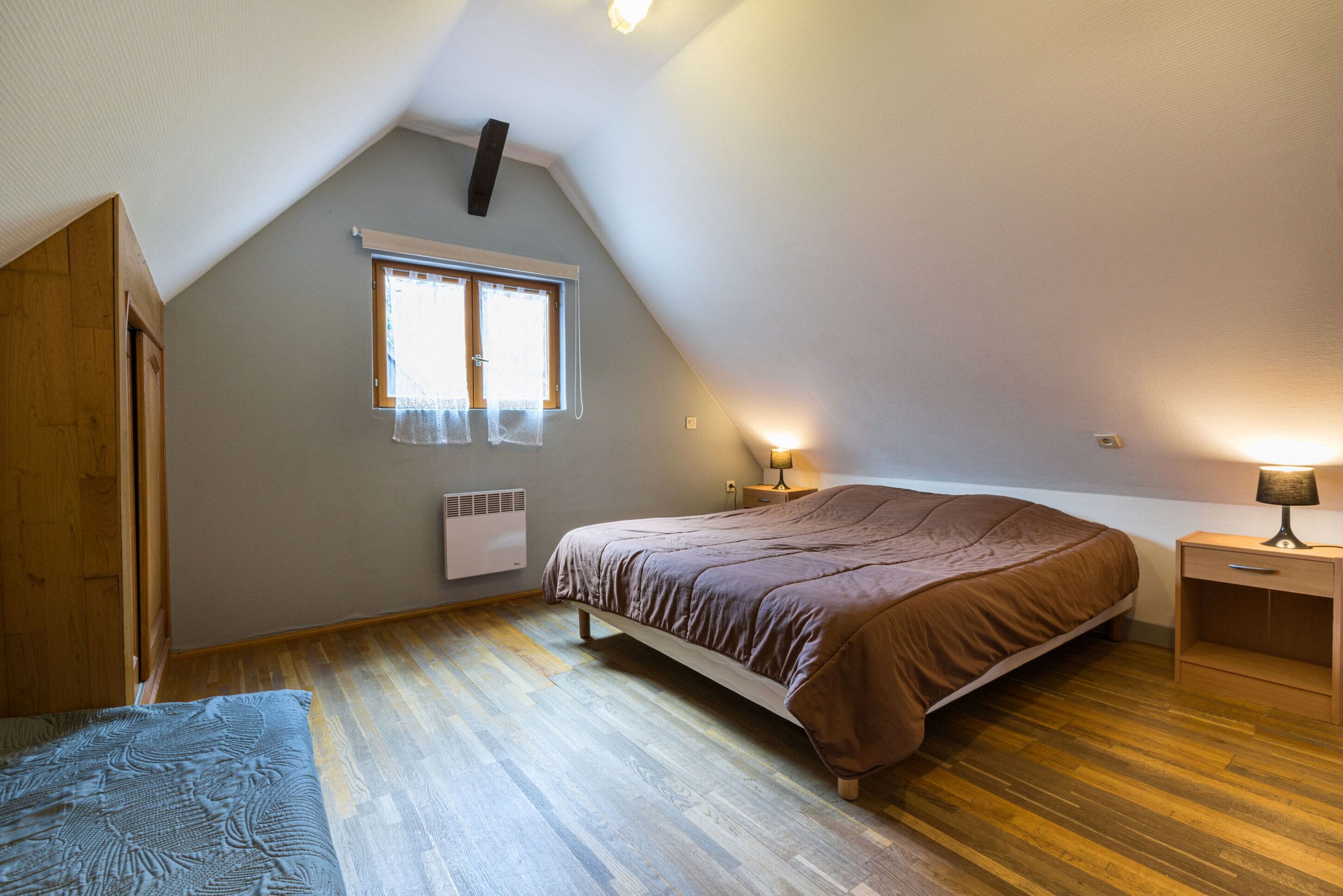 A bedroom with a sloped ceiling, a bed, a window with curtains, a bedside table with a lamp, and a radiator.