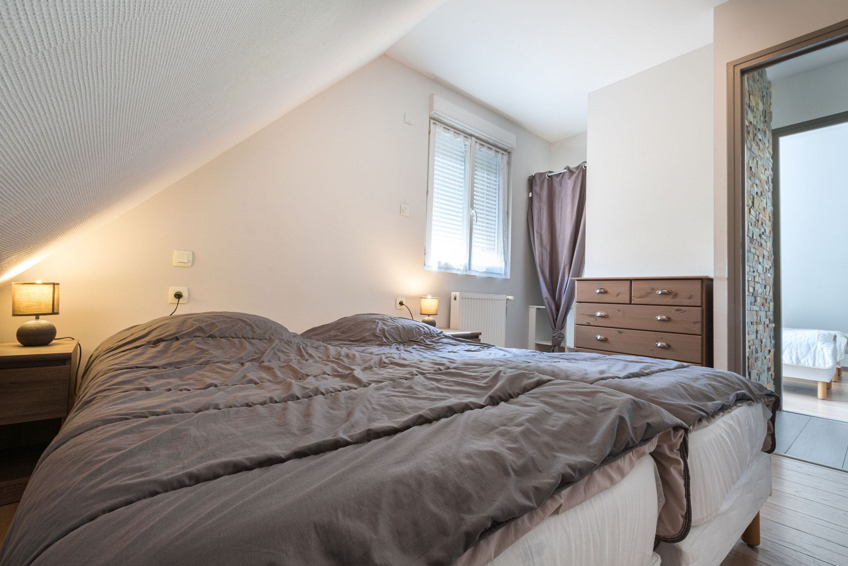 A bedroom with a large bed, a dresser, a nightstand, a window with blinds, and a radiator.