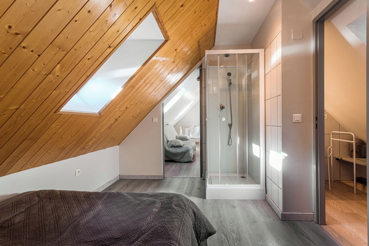 Un dormitorio con techo inclinado, cabina de ducha de vidrio y cama con manta oscura.