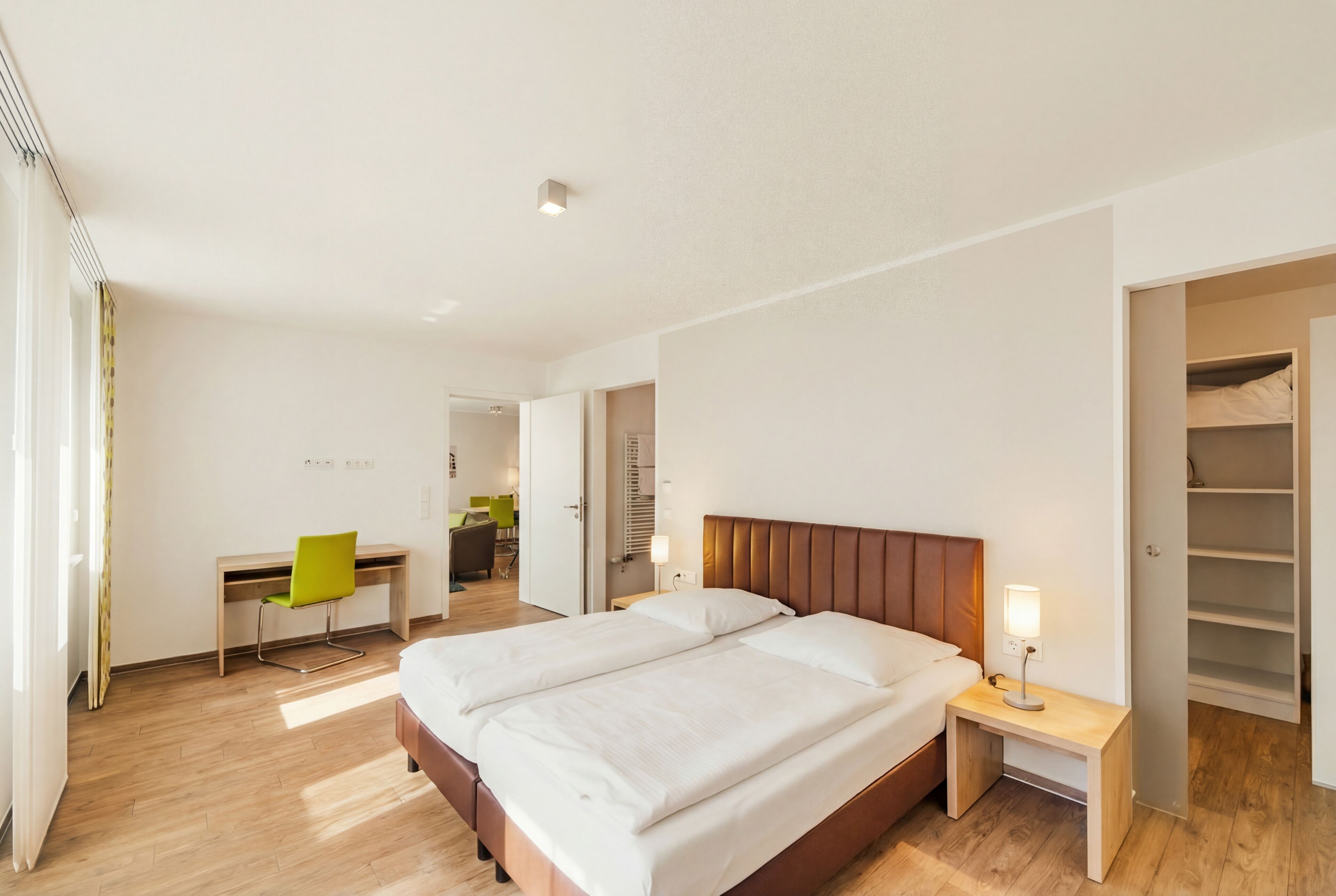 A modern hotel room with a double bed, a desk with a chair, a wardrobe, and a bathroom visible in the background.