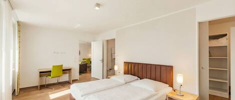A modern hotel room with a double bed, a desk with a chair, a wardrobe, and a bathroom visible in the background.