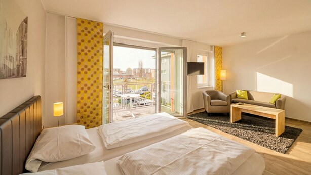 A hotel room with a bed, a sofa, a TV, and a balcony with a view of the city. - ADAPT APARTMENT HOTEL Berlin-Adlershof (Berlin)