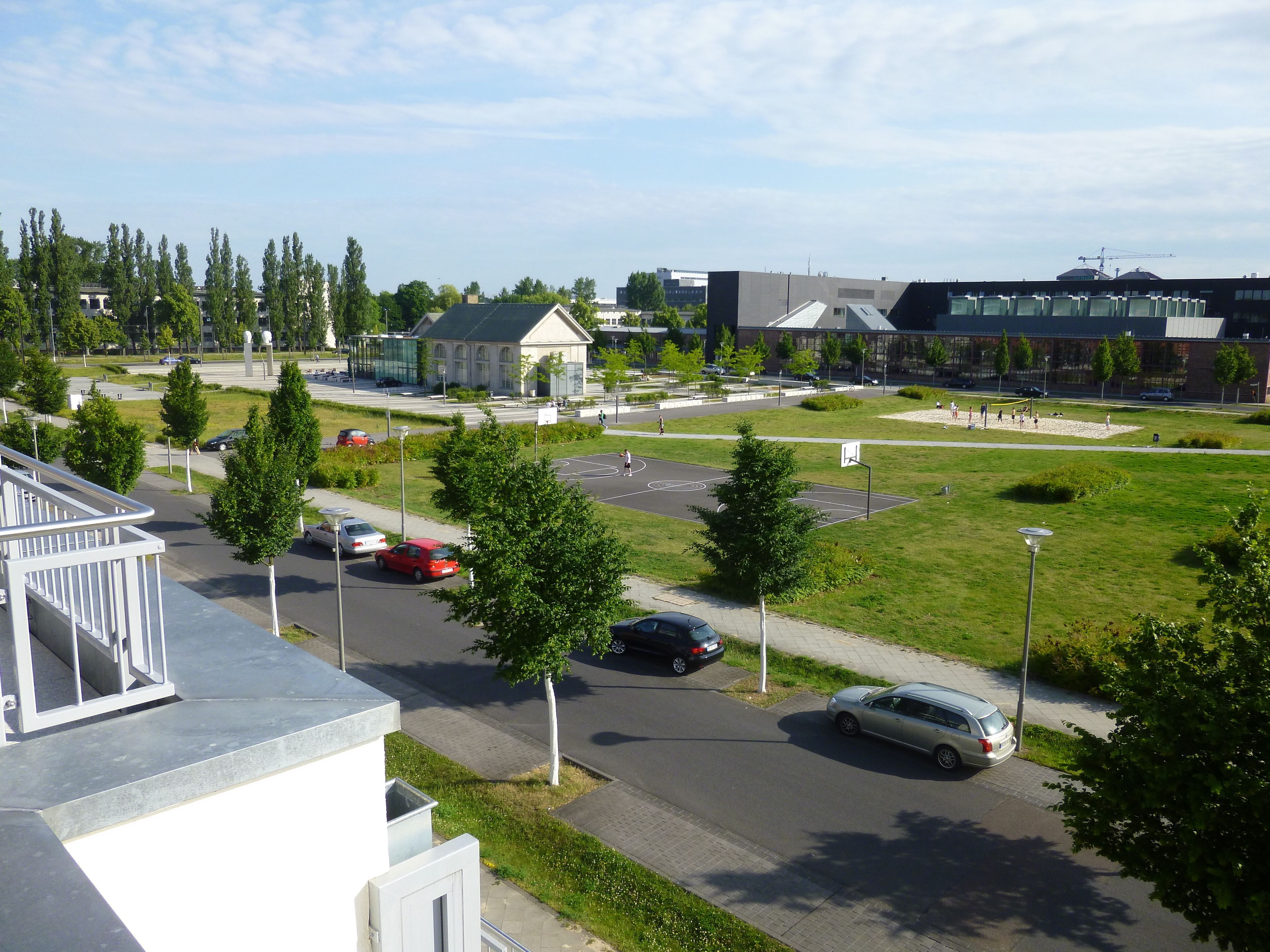 A parking lot with cars, a basketball court, and a building with a green roof.