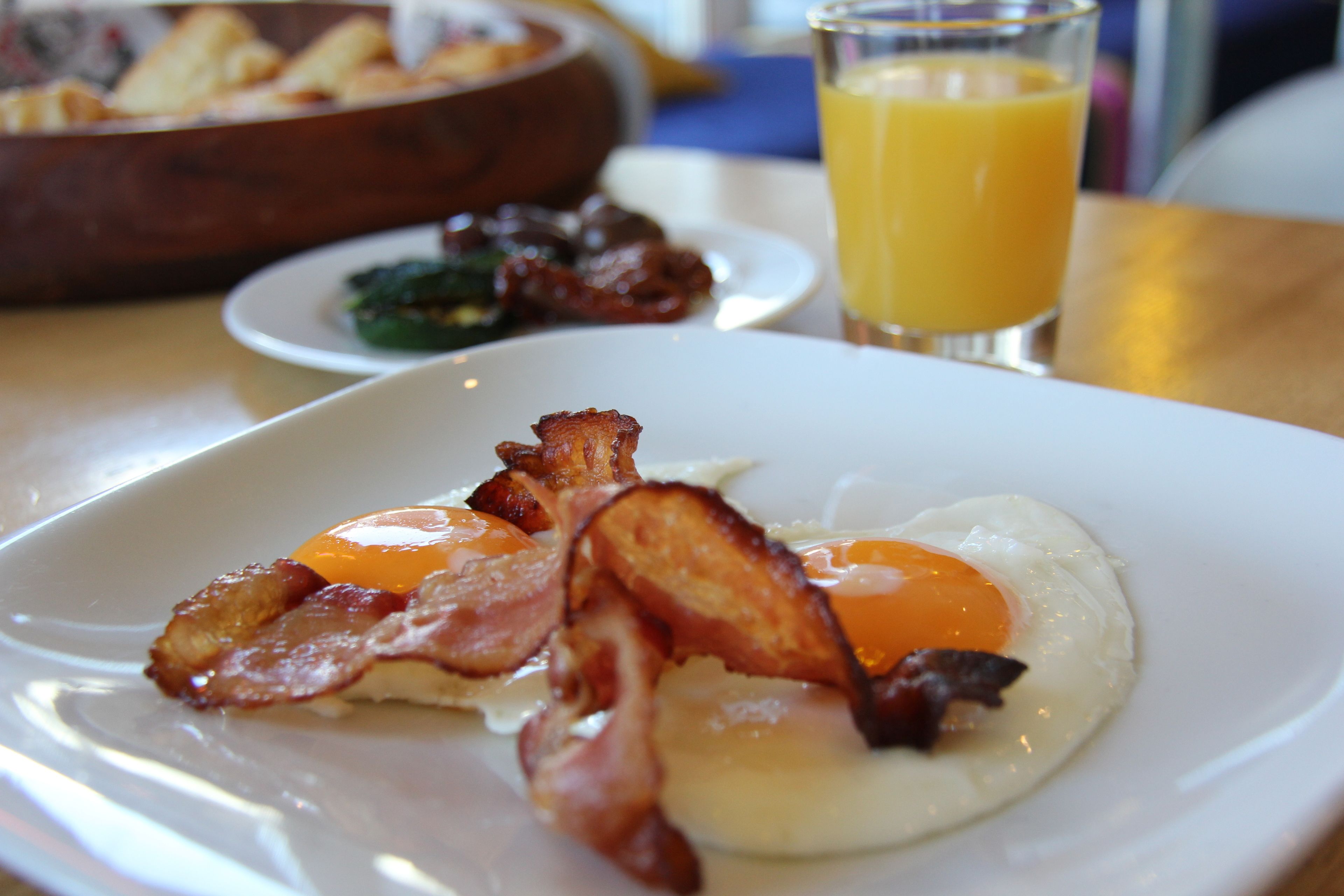 A breakfast plate with eggs and bacon, a glass of orange juice, and a bowl of toast in the background.