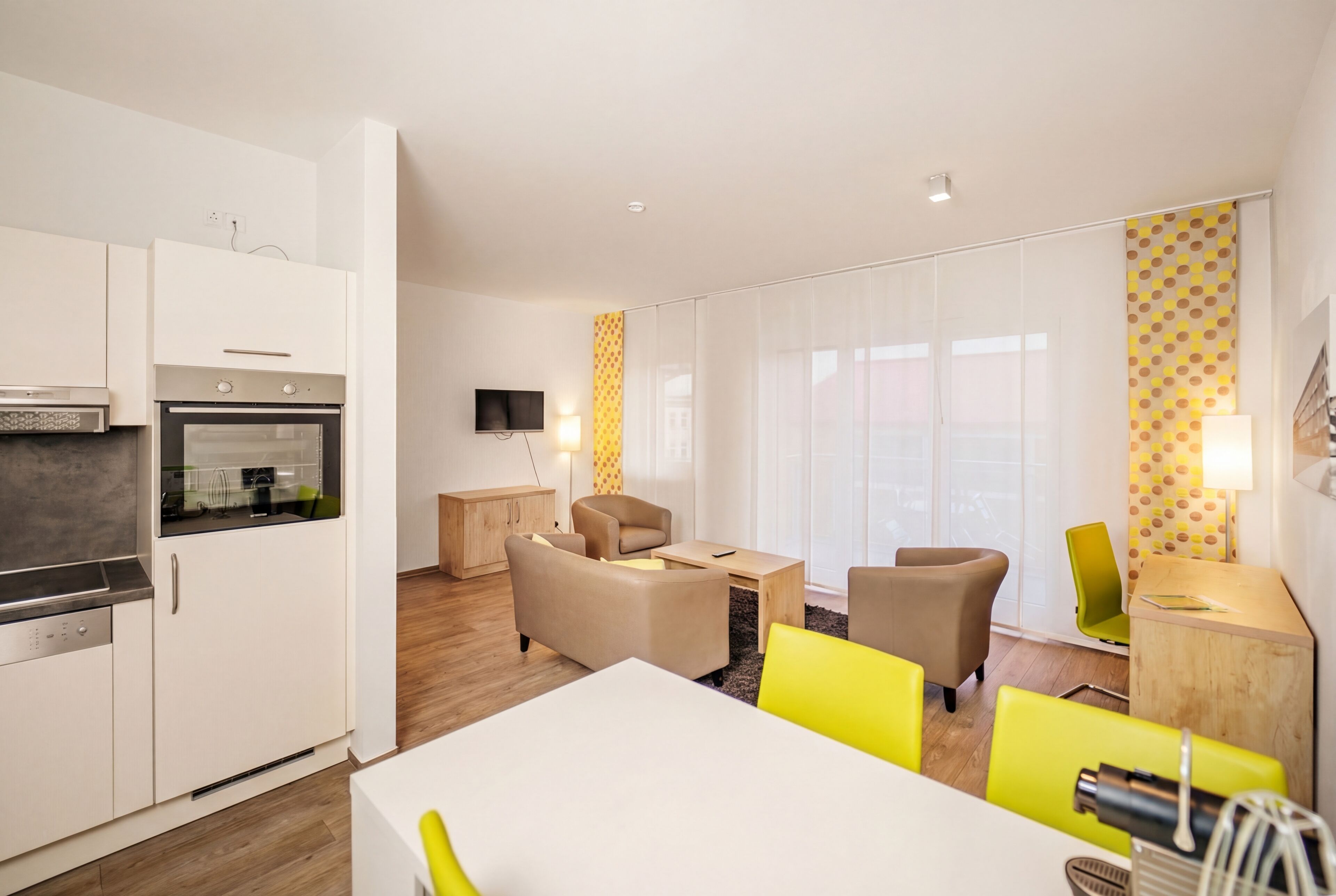 A modern kitchen with built-in appliances, a dining area with yellow chairs, and a living space with a TV and large windows.