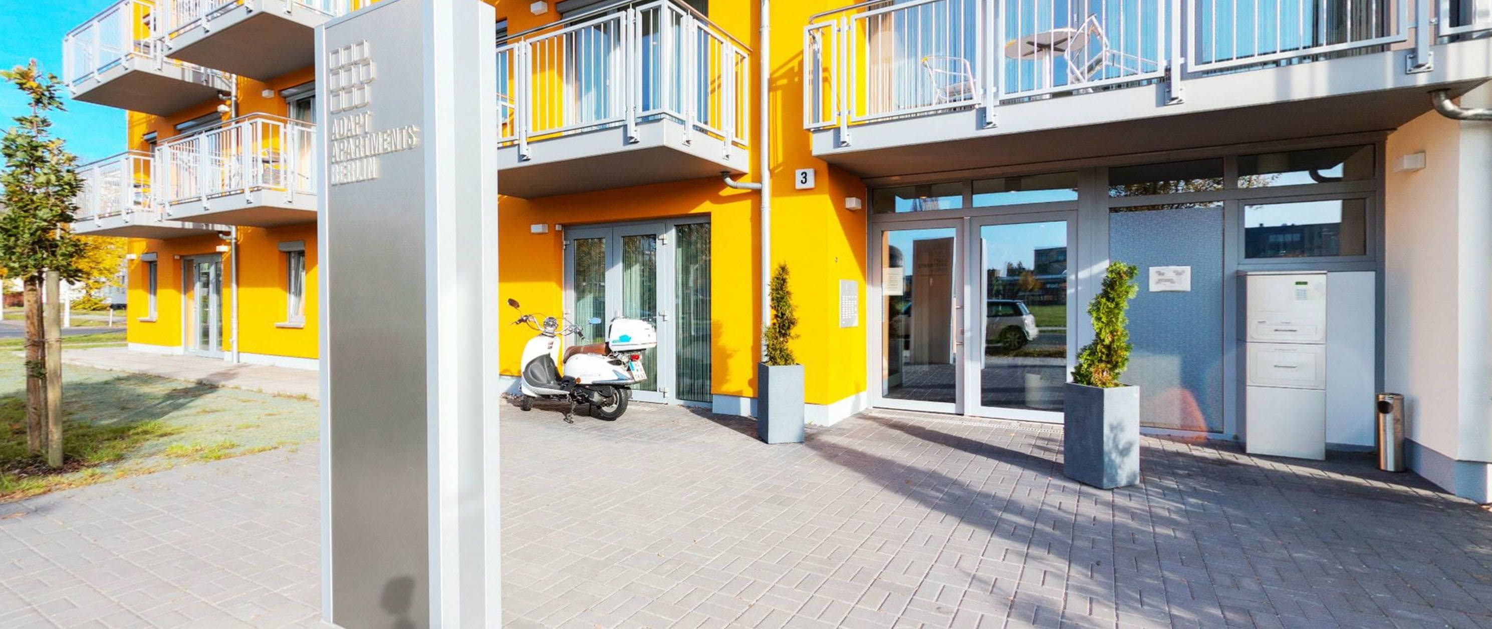 A multi-story yellow building with balconies, a scooter parked in front, and a clear sky.