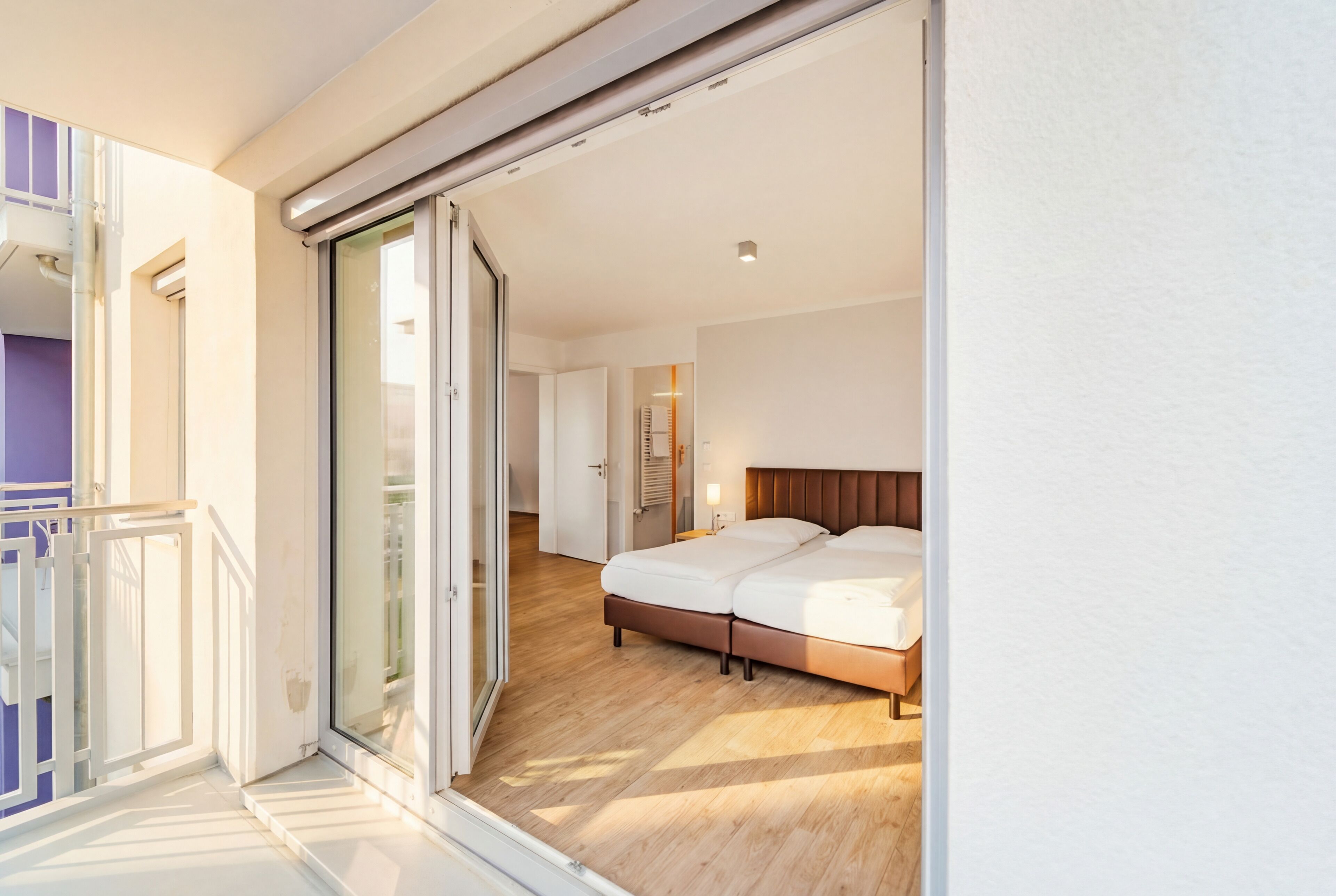 A modern hotel room with a sliding glass door leading to a balcony, a large bed, and a wooden floor.