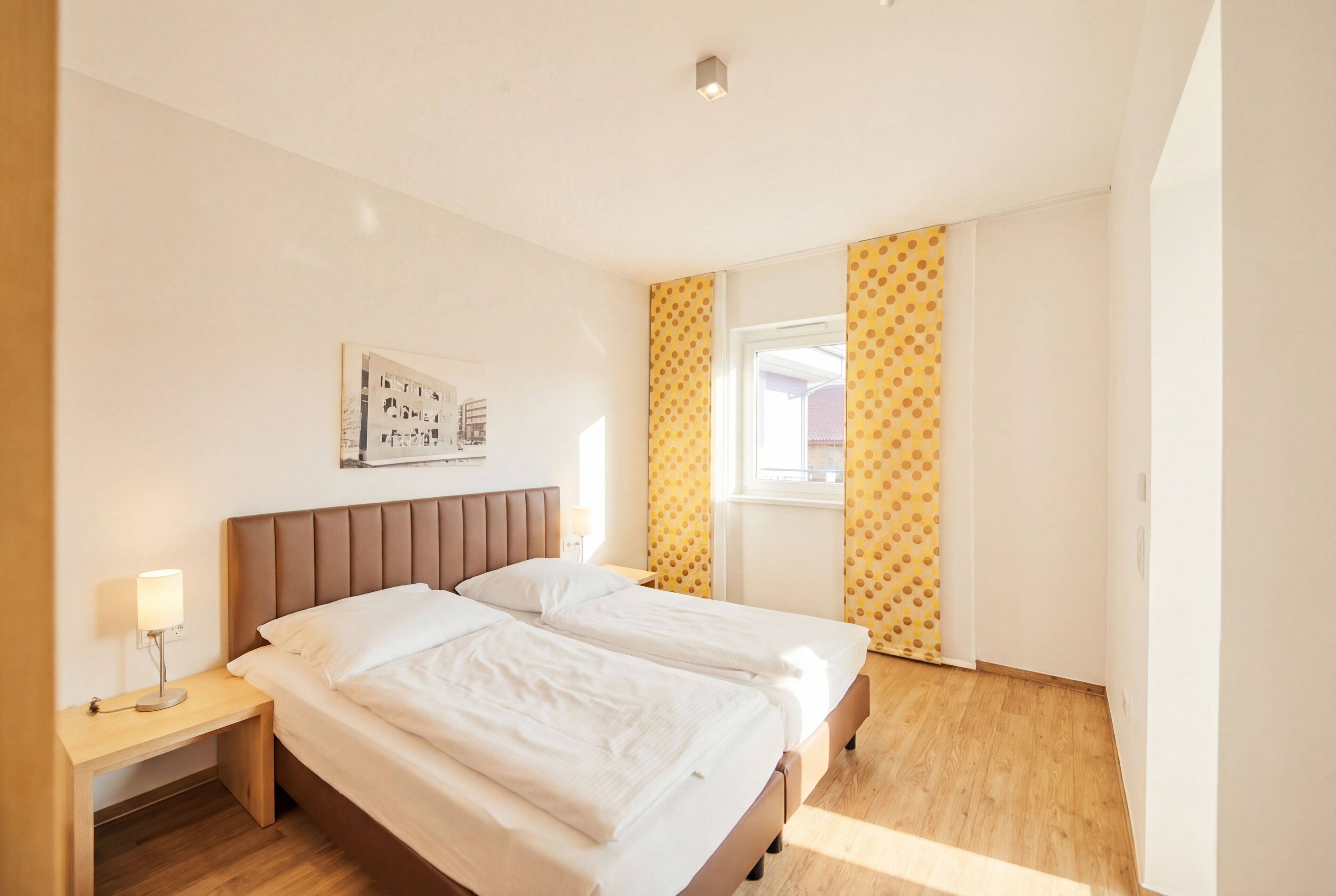 A hotel room with two beds, a nightstand with a lamp, a window with yellow curtains, and a wall-mounted picture.