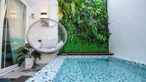 Una piscina interior moderna con diseño en mosaicos, una silla colgante y una pared verde exuberante con diversas plantas.