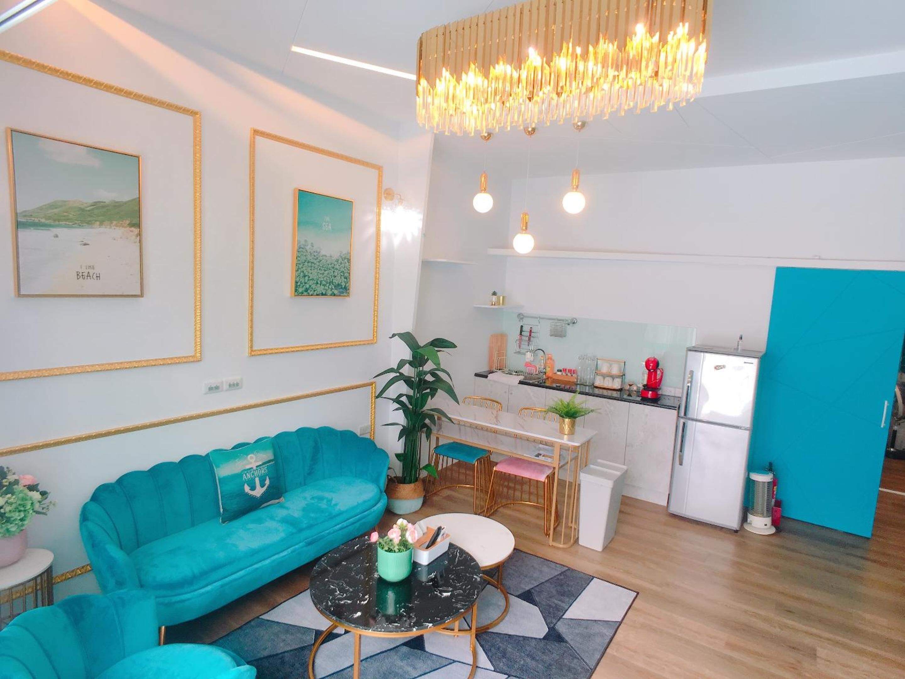 A modern living room with a teal sofa, a dining area with a small table, and a kitchen with a white refrigerator and a blue accent wall.