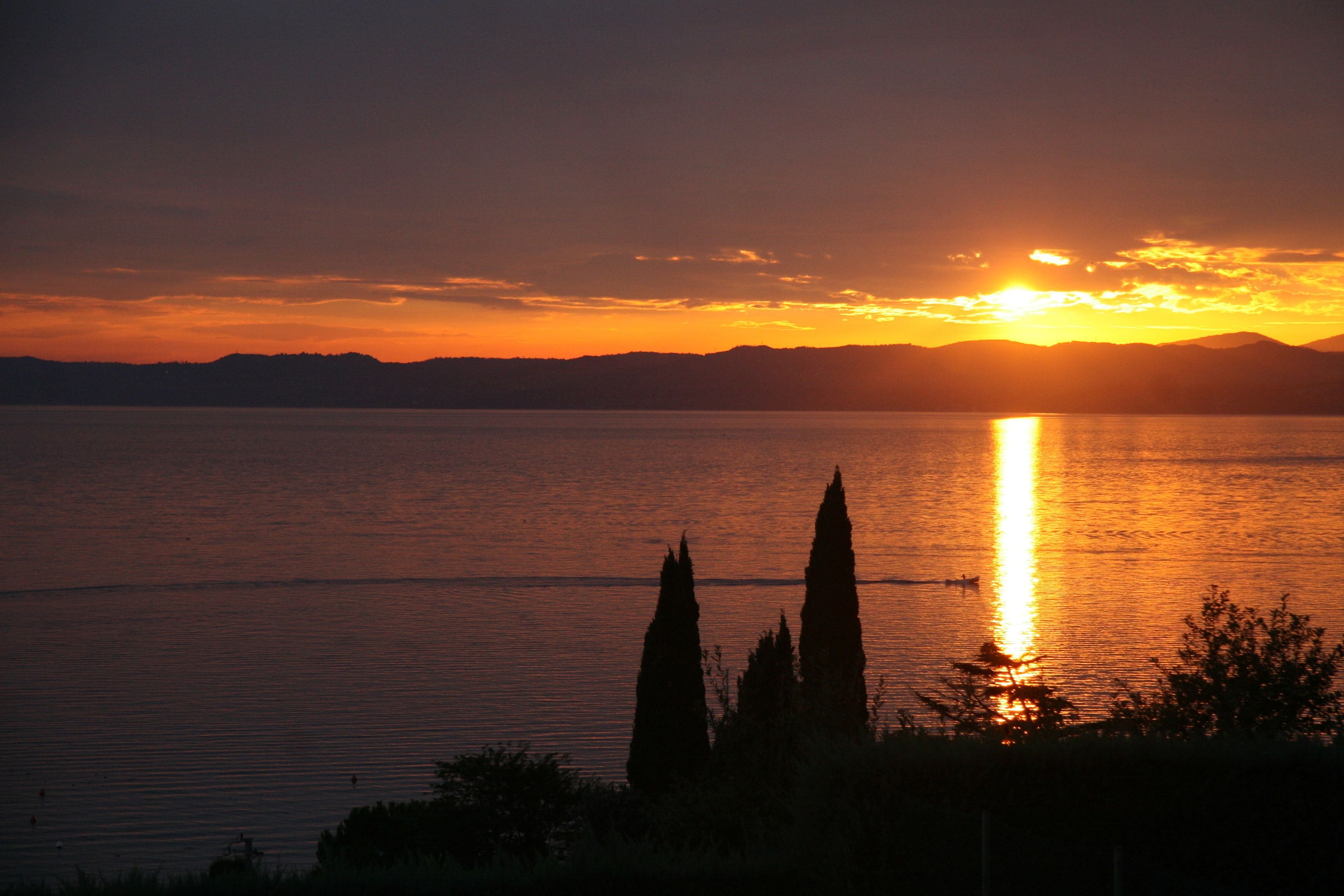 Un coucher de soleil sur un lac paisible, avec les silhouettes d’arbres et de montagnes lointaines.
