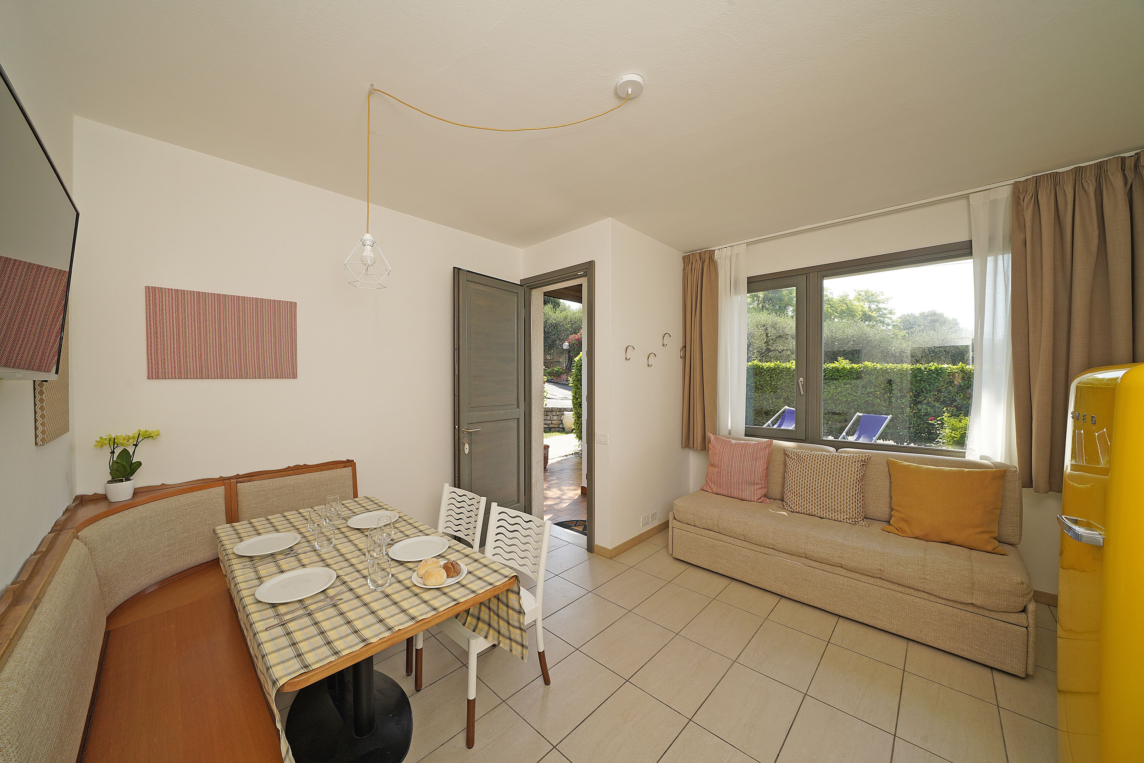 A dining area with a table set for four, a sofa with cushions, a potted plant, and a view of the outdoors through a window.