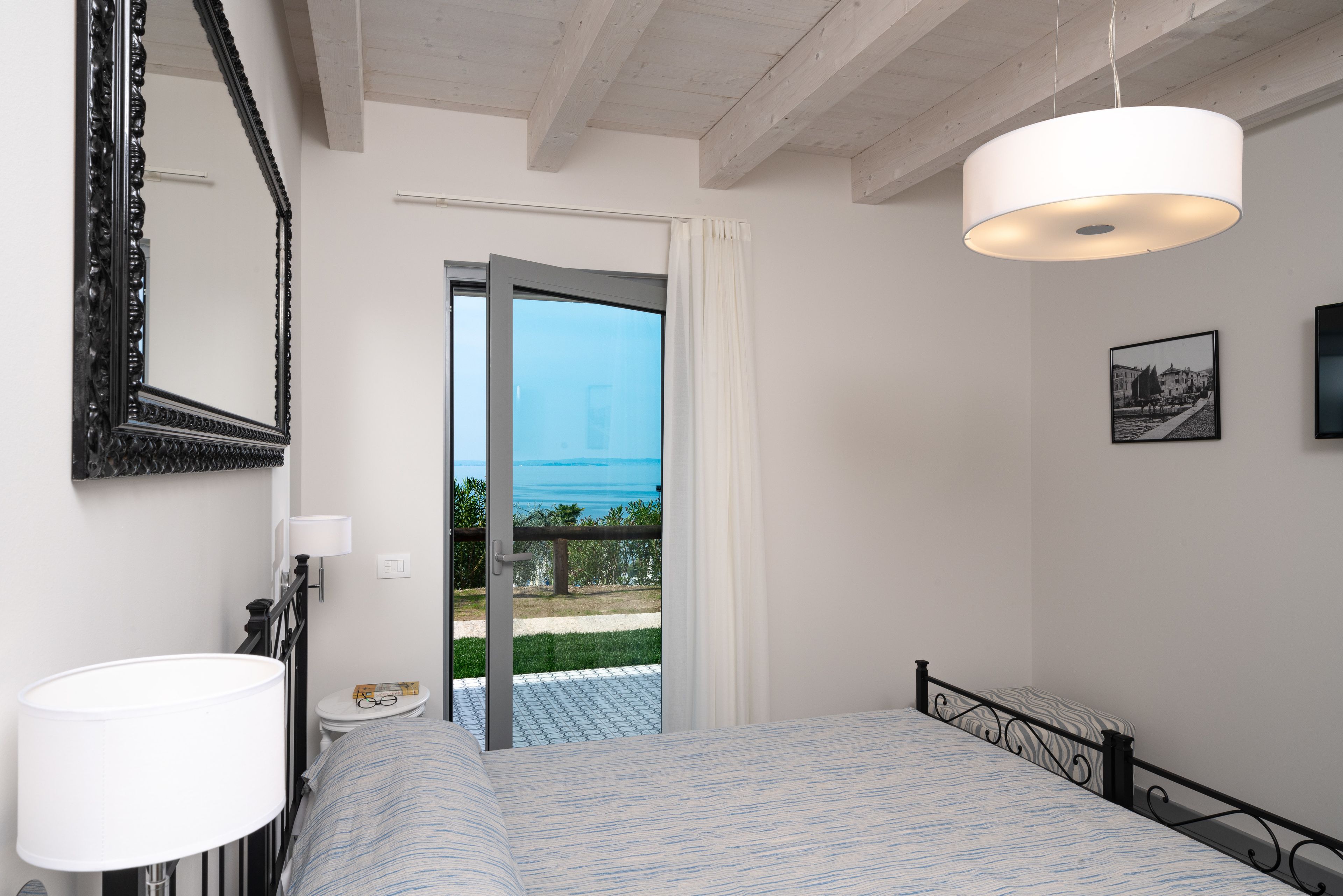 A bedroom with a bed, a bedside table, a lamp, a mirror, and a door leading to a balcony with a view of the sea.