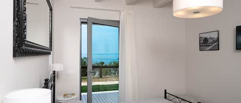 A bedroom with a bed, a bedside table, a lamp, a mirror, and a door leading to a balcony with a view of the sea.