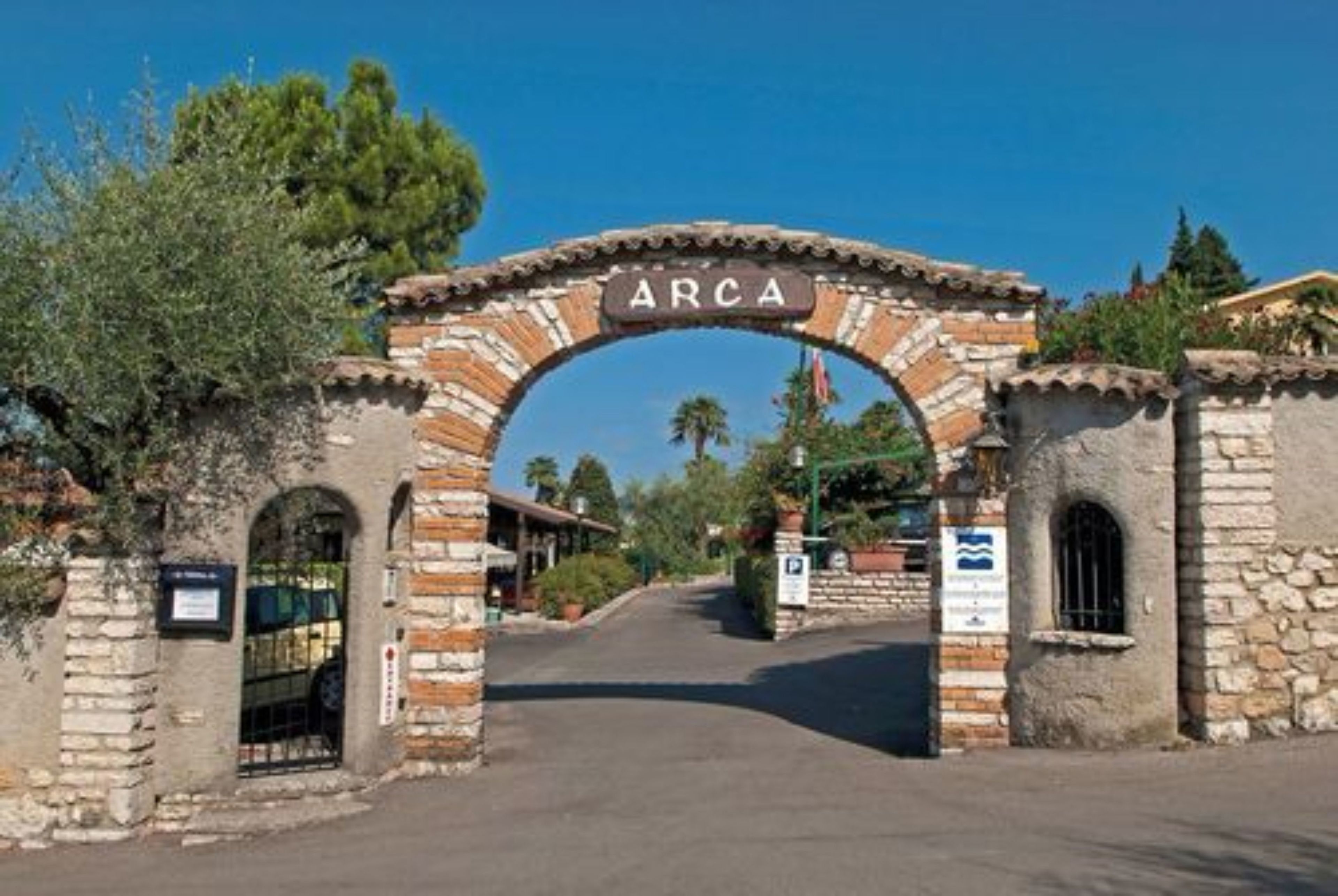 An arched stone gateway with the word "ARCA" inscribed above it, leading to a street with buildings and greenery.