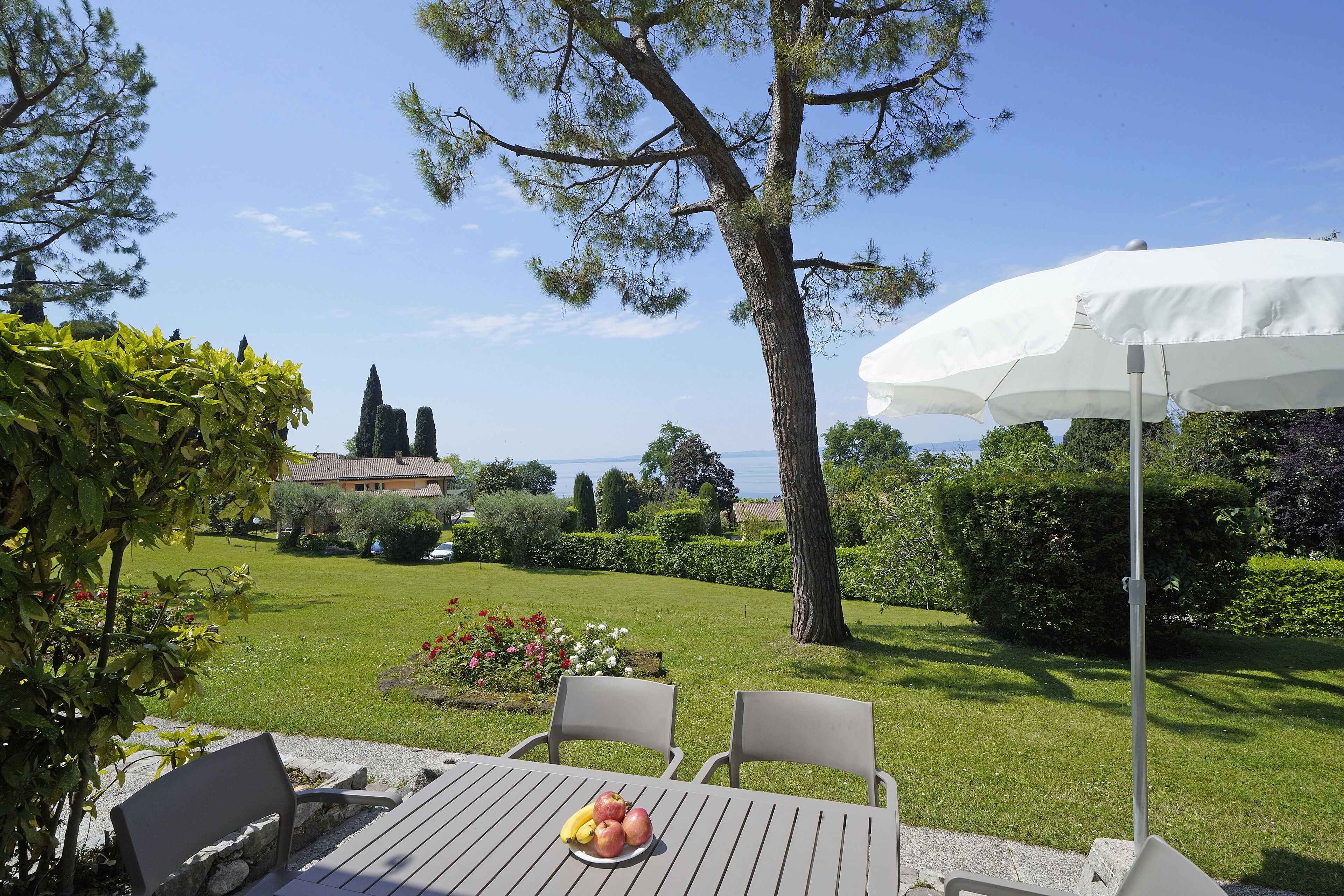 Eine Terrasse mit Tisch für zwei Personen, einem weißen Sonnenschirm und Blick auf einen gepflegten Garten sowie entfernte Gebäude.