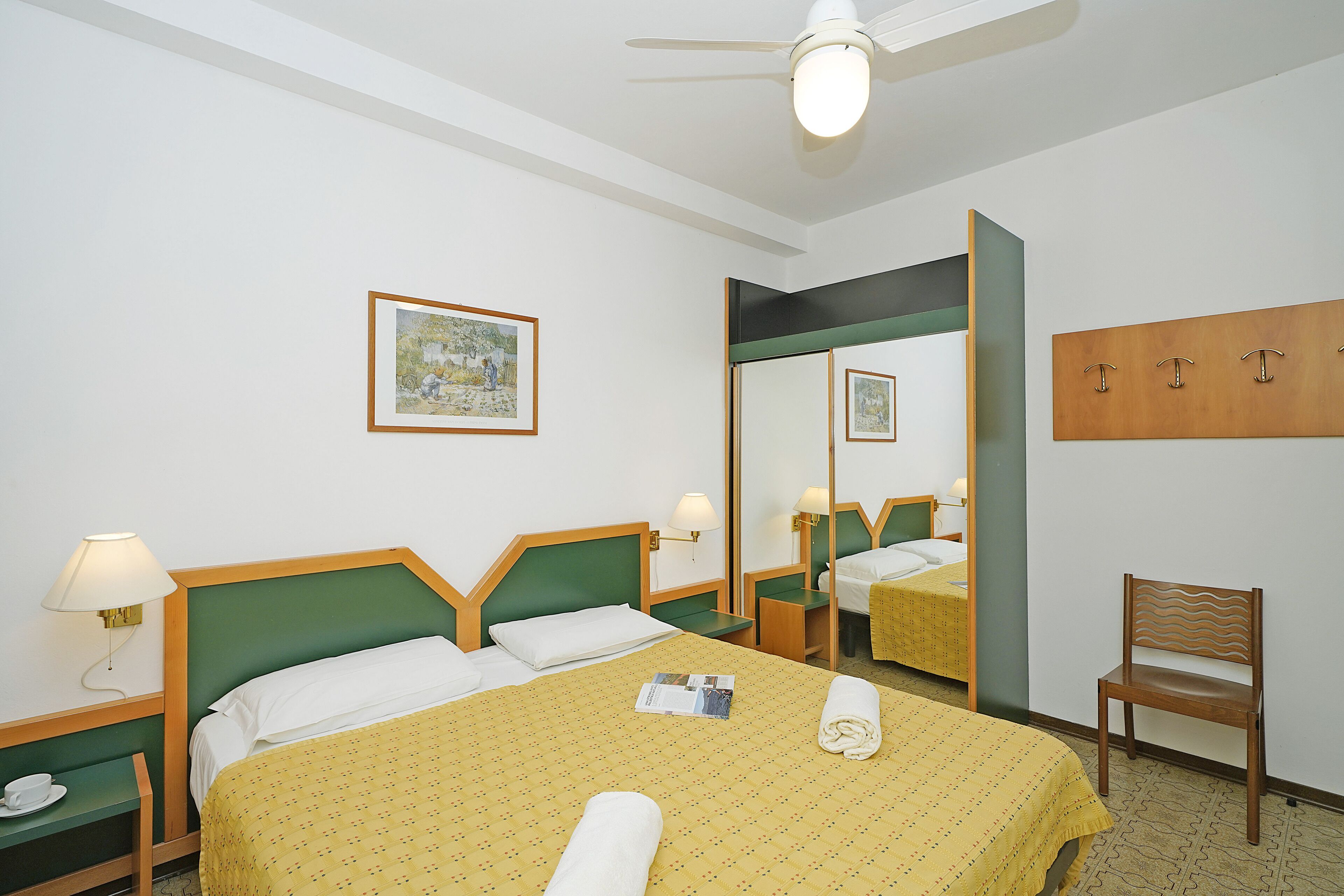 A hotel room with a bed, a chair, a nightstand, a lamp, a mirror, and a wall-mounted coat rack.