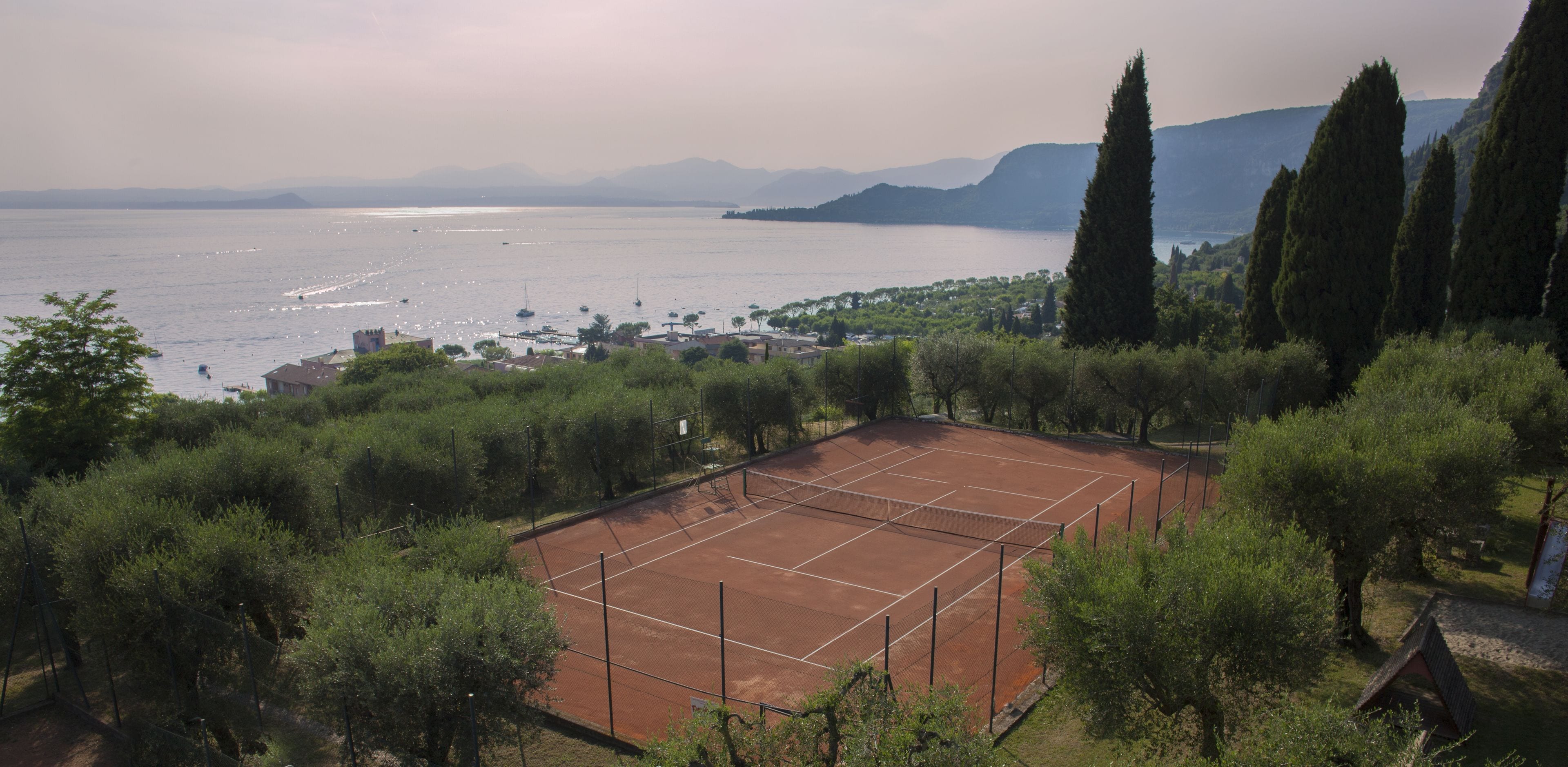 A tennis court surrounded by trees, with a view of the sea and mountains in the background.