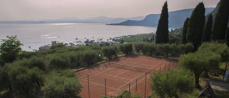 A tennis court surrounded by trees, with a view of the sea and mountains in the background.