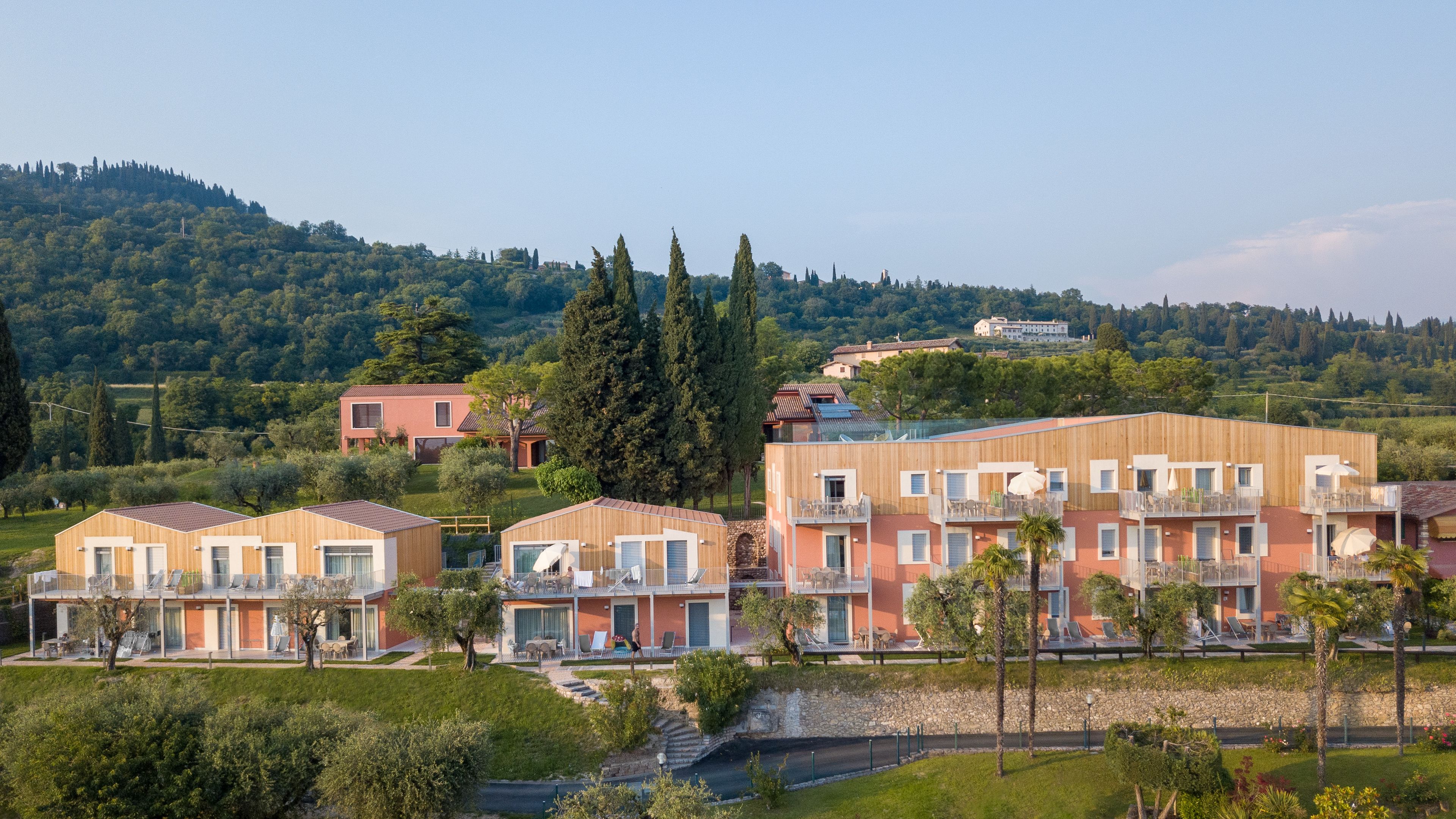 A modern residential complex with multiple two-story buildings, featuring balconies with outdoor furniture and a lush green landscape in the background.
