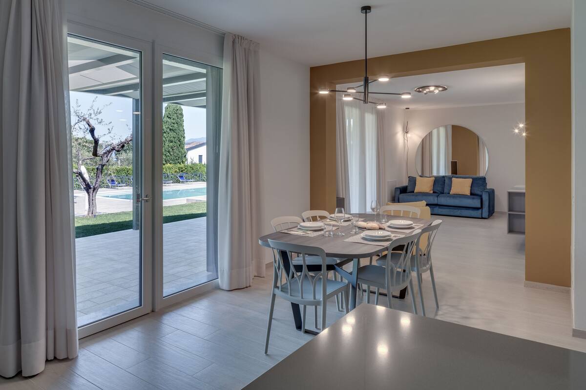 Un espace de repas moderne avec une table dressée pour quatre personnes, un canapé en arrière-plan et une vue sur une piscine à travers des portes vitrées.