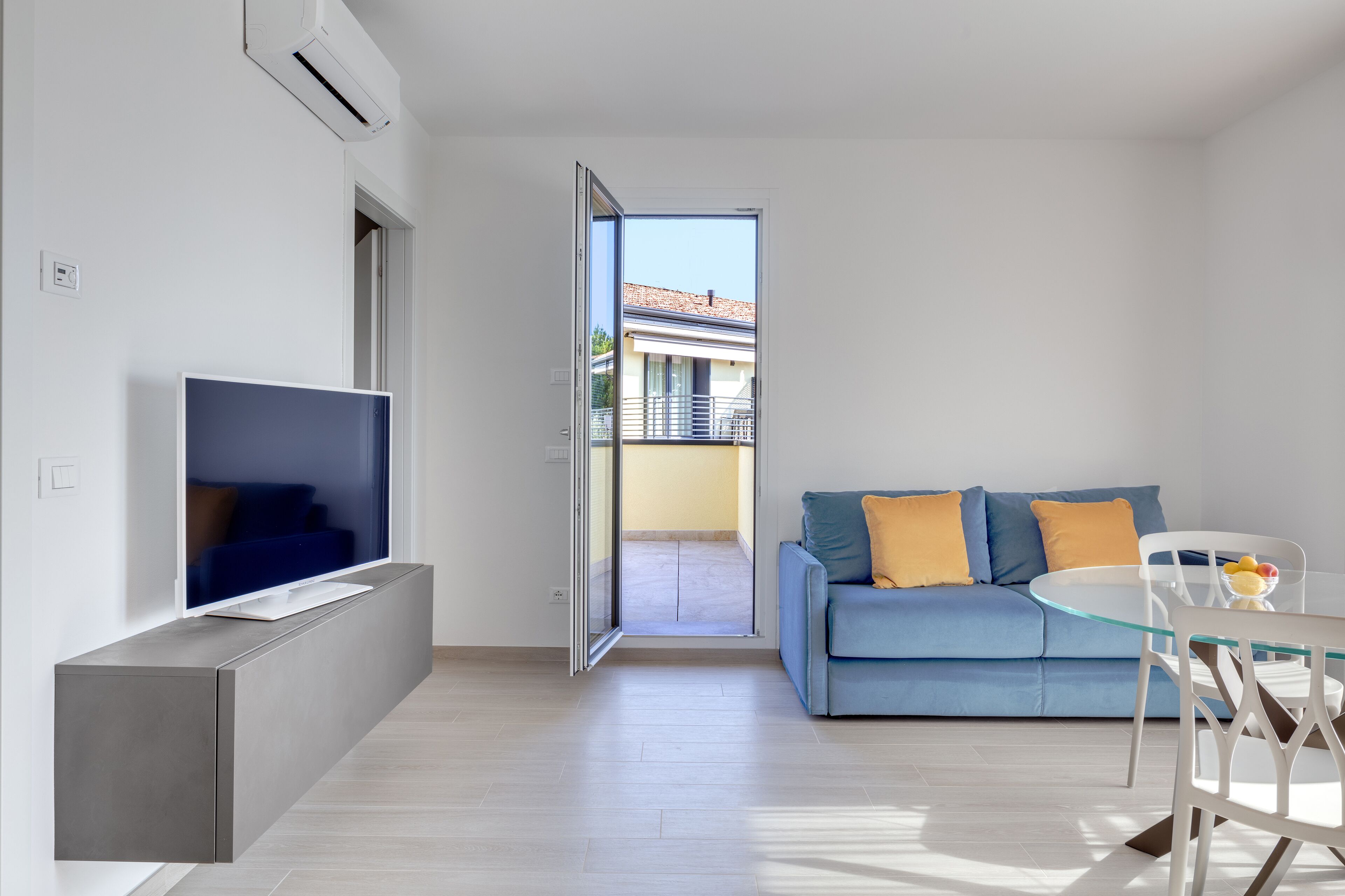 Una sala moderna con un televisor de pantalla plana, un sofá azul con cojines amarillos, una mesa de comedor de vidrio y un ventanal corredizo que da a un balcón exterior.
