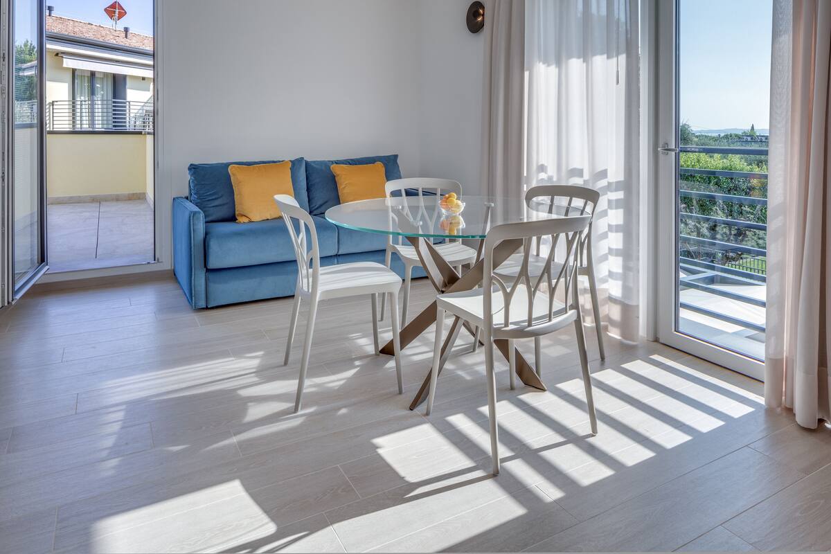 Un salon moderne avec une table en verre et des chaises blanches, un canapé bleu avec des coussins jaunes, et une vue sur un balcon et un immeuble à travers de grandes fenêtres.