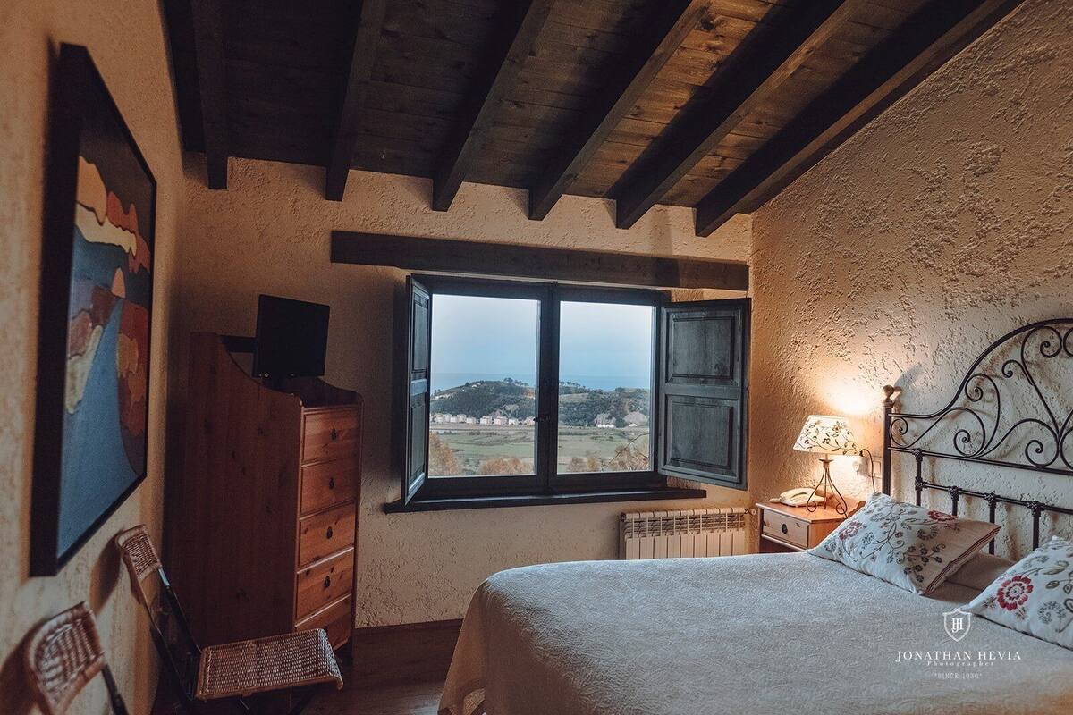 A bedroom with a bed, a dresser, a chair, a window with a view of the countryside, and a lamp on a bedside table.