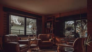A cozy living room with leather chairs, a small round table, and a bookshelf. Large windows offer views of a garden and a wooden fence. - Paraje del Asturcón Adults Only (Ribadesella)