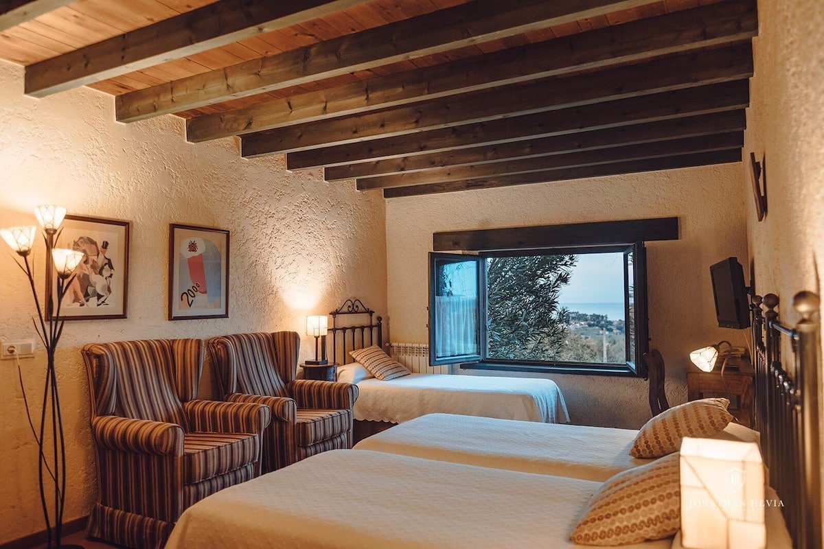 A bedroom with two beds, a striped armchair, a bedside table with a lamp, a framed picture on the wall, and a window with a view.