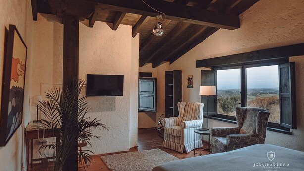 A spacious room with a high ceiling, a large window offering a scenic view, a television mounted on the wall, and a comfortable seating area with a patterned armchair and a sofa. - Paraje del Asturcón Adults Only (Ribadesella)