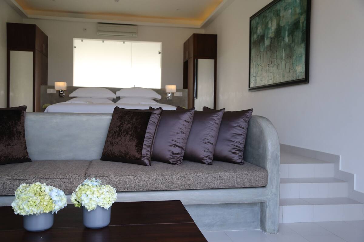 A modern bedroom with a grey sofa, a bed with white linens, and a large window. There are two bedside lamps and a painting on the wall.