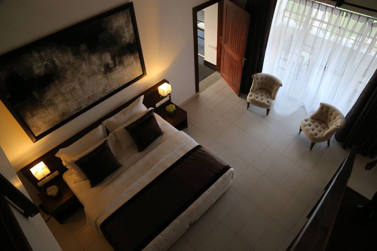 A hotel room with a large bed, two bedside tables with lamps, a large abstract painting on the wall, and two armchairs near a window with sheer curtains.