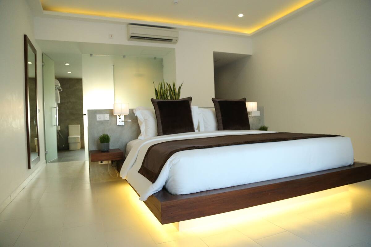 A modern hotel room with a large bed, a bedside table with a lamp, a potted plant, and a mirror.