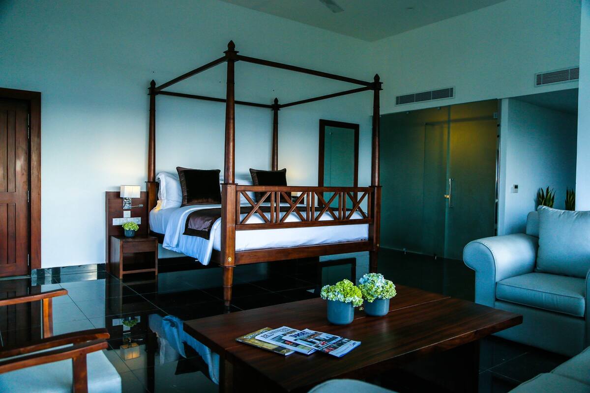 A four-poster bed with a wooden frame, a small bedside table with a lamp, a sofa, and two potted plants on a wooden table.