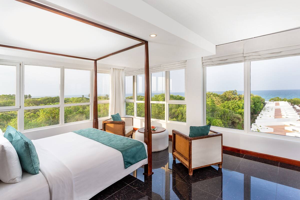 Een moderne hotelkamer met een groot bed, een zithoek met een stoel en een tafeltje, en uitzicht op zee en groen.