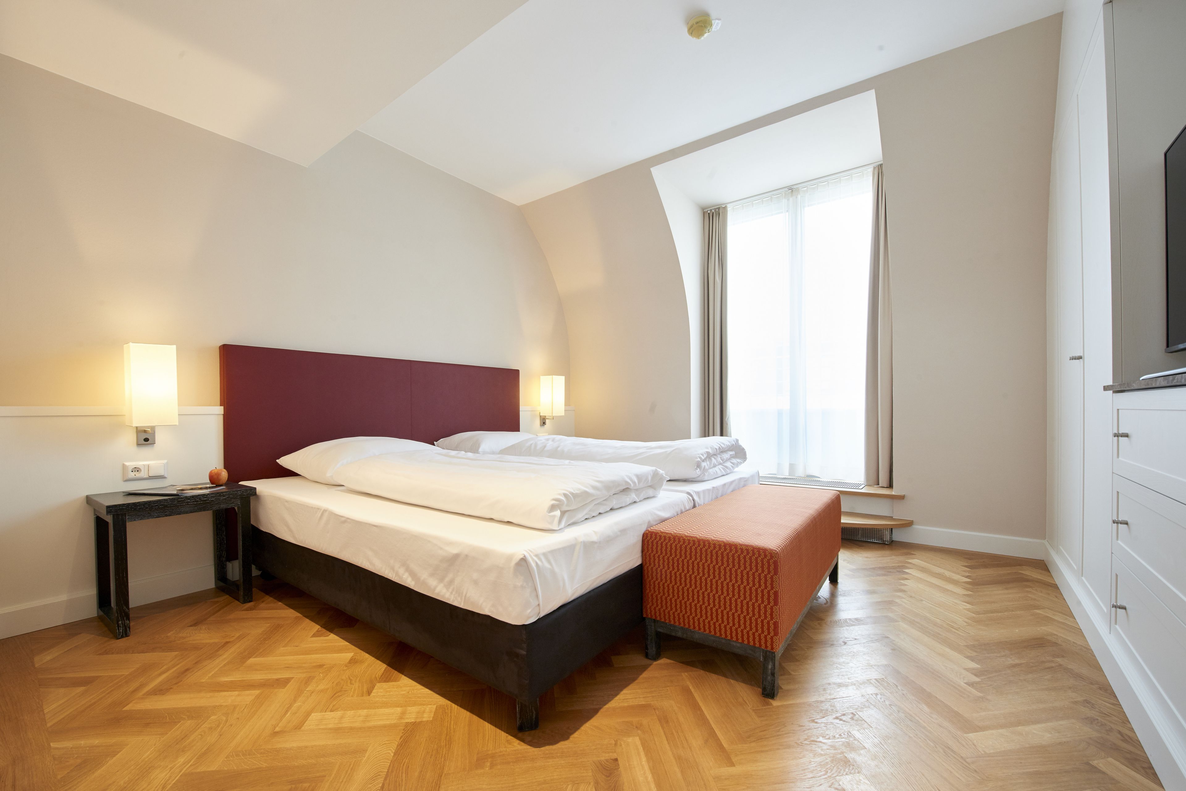 A hotel room with a bed, bedside tables, a bench, a TV, and a window with curtains.