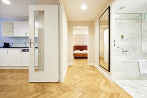 Quarto de hotel moderno com piso de madeira, chuveiro de vidro e cama com cabeceira vermelha.