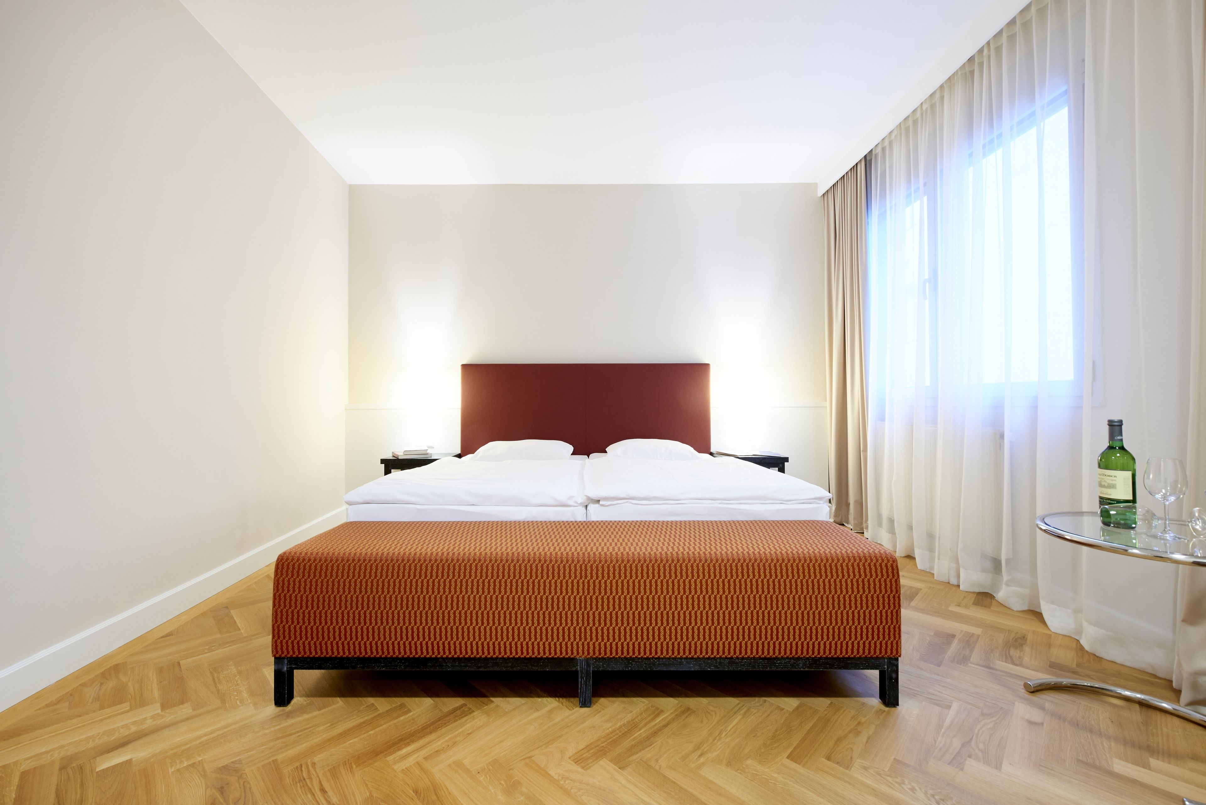 A hotel room with a bed, a nightstand, a glass table with a bottle, and a window with curtains.